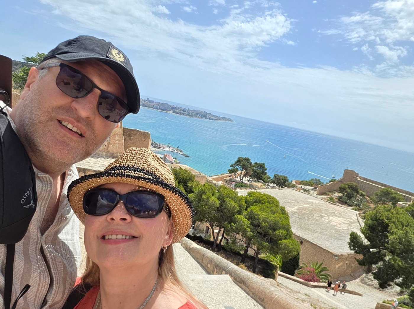 The parents, smiling in a selfie at a hilltop fortress above a blue sea, both in sunglasses and hats